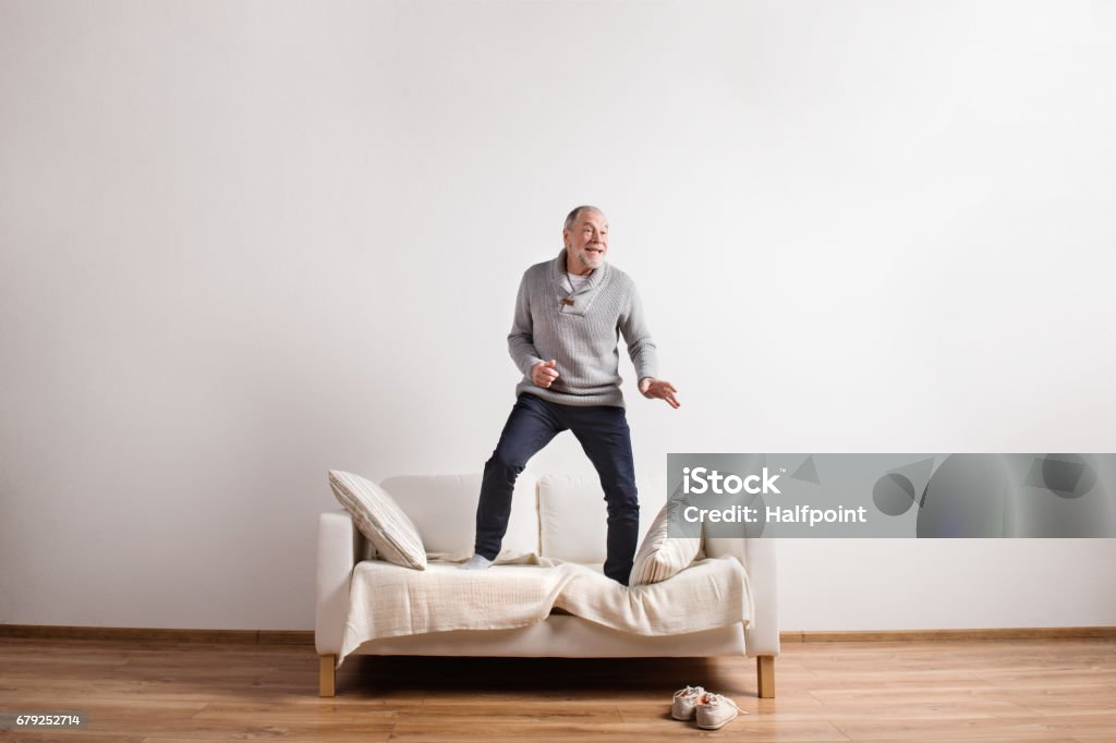 Handsome senior man standing on couch, dancing. Studio shot. Handsome senior man standing on couch, dancing, having fun. Studio shot against white wall. Sofa Stock Photo