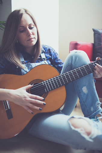 Young hipster girl playing guitar at cozy home interior , pretty woman learning to play guitar at home.