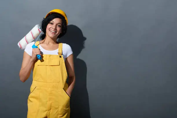 Happy smiling mixed race construction woman wearing yellow protect helmet and overall adjusting helmet holding a painting roller over shoulder standing at grey wall looking at camera.