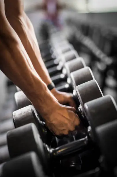 Close up of man hands holding dumbbells at gym. Strong bodybuilder lifting dumbbell. Detail of guy hands taking dumbbells from row of barbells in a gym.