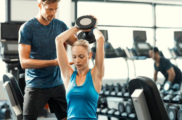 Woman lifting dumbbells Trainer helping athletic woman at gym. Personal trainer giving weightlifting training to girl in gym. Young woman working out at gym using dumbbells with help of personal trainer. fitness instructor stock pictures, royalty-free photos & images