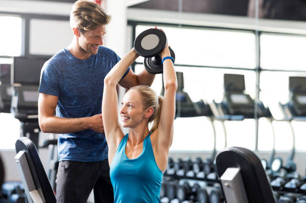 Woman with personal trainer Smiling woman using dumbbells with personal trainer in the fitness room. Young woman doing weight exercises with assistance of man in gym. Personal coach helping happy girl to do exercises with dumbbells. fitness trainer stock pictures, royalty-free photos & images