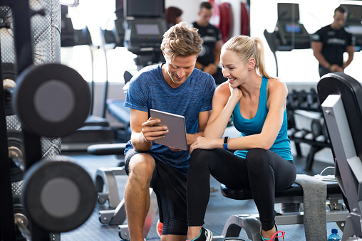 Smiling personal trainer using digital tablet while talking to blonde woman at gym. Happy couple using tablet in fitness club. Client trainer looking at computer her progress at the gym.