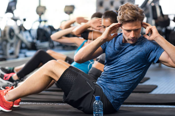 Man doing sit ups Muscular guy doing sit ups at gym with other people in background. Young athlete doing stomach workout in modern gym. Handsome fit man doing crunches at gym. sit ups stock pictures, royalty-free photos & images