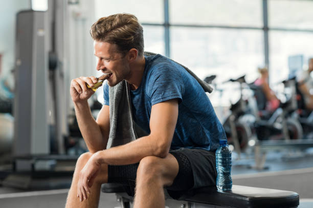 Man eating energy bar Sweaty young man eating energy bar at gym. Handsome mid guy enjoying chocolate after a heavy workout in fitness studio. Fit man biting a snack and resting on bench. sportsperson stock pictures, royalty-free photos & images