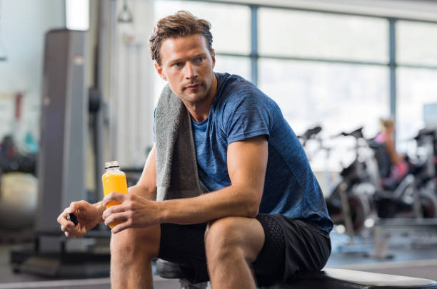 Fit man with energy drink Handsome young man in sportswear holding bottle of fresh orange juice while resting at gym.Thoughtful fit man sitting alone holding a bottle of energy drink. Guy take break after fitness exercise on bench. sport drink stock pictures, royalty-free photos & images