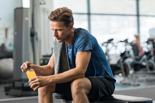 Young man in gym sitting alone opening a bottle of energy drink. Thoughtful fit man in gym holding fruit juice and thinking. Tired man in gym resting while drinking fruit juice.