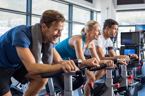 Group of smiling friends at gym exercising on stationary bike. Happy cheerful athletes training on exercise bike. Young men and woman working out at exercising class in the gym.