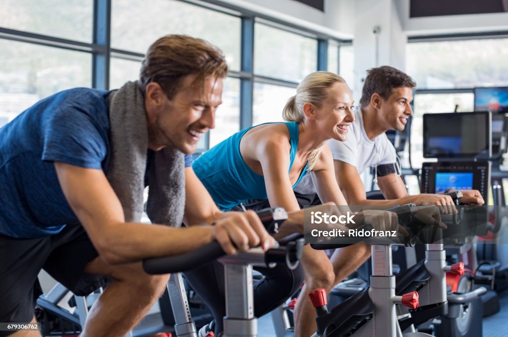 People on exercise bike Group of smiling friends at gym exercising on stationary bike. Happy cheerful athletes training on exercise bike. Young men and woman working out at exercising class in the gym. Stationary Cycling Class Stock Photo