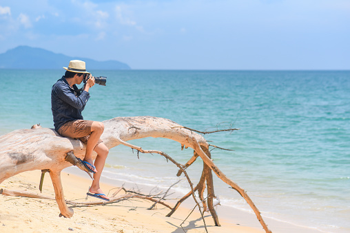 young man photographer with jean shirt and hat sitting on white dead tree near sandy beach of tropical island and take photos of turquoise sea, summer holiday and vacation travel concepts