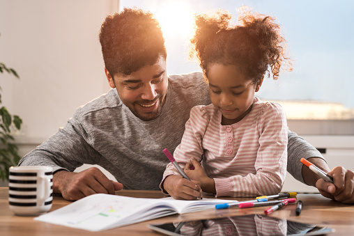 Happy African American father and daughter coloring on a piece of paper while spending their free time at home.