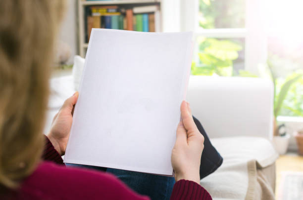 Blond woman sitting in a living room at home holding a blank brochure with copy space in hands. stock photo