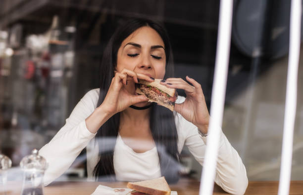 bela mulher sorridente jovem comendo sanduíche no café. janela de café - sandwich healthy eating wrap sandwich food - fotografias e filmes do acervo