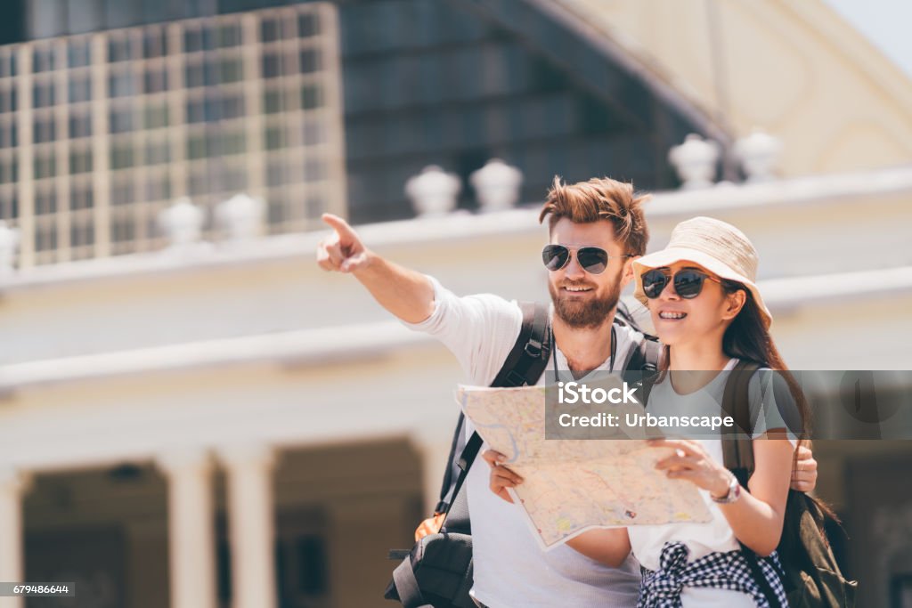Multiethnic traveler couple using generic local map together on sunny day, man pointing toward copy space. Honeymoon trip, backpacker tourist, Asia tourism, or holiday vacation travel concept Tourist Stock Photo