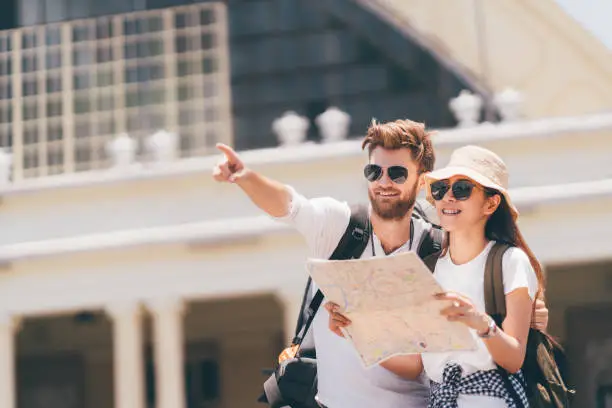 Photo of Multiethnic traveler couple using generic local map together on sunny day, man pointing toward copy space. Honeymoon trip, backpacker tourist, Asia tourism, or holiday vacation travel concept