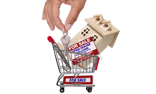 Hand dropping key in shopping cart with wood house and for sale open house real estate sign isolated on white background