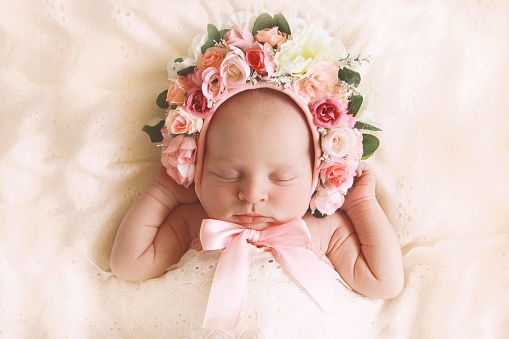 A newborn baby girl, fast asleep, wears a bonnet of flowers.