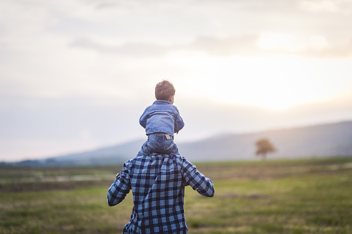 Father carries his baby boy on the shoulder while they walk through the field in the nature