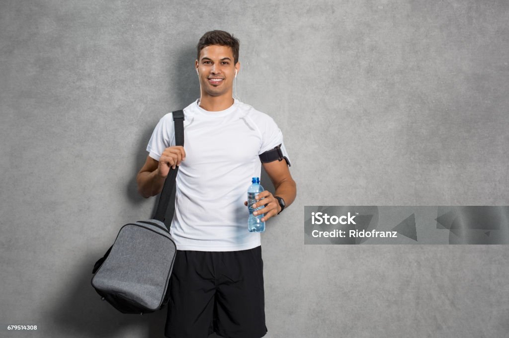 Man with gym bag Portrait of young man athlete with bag and bottle of water. Fitness guy isolated over grey background looking at camera. Latin man listening to music after workout and leaning against grey wall. Men Stock Photo