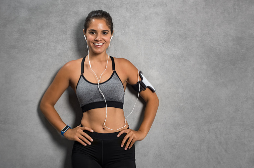 Happy young fitness woman with armband standing and listening to music over grey background. Beautiful smiling girl wearing sportswear and looking at camera leaning on wall with copy space.