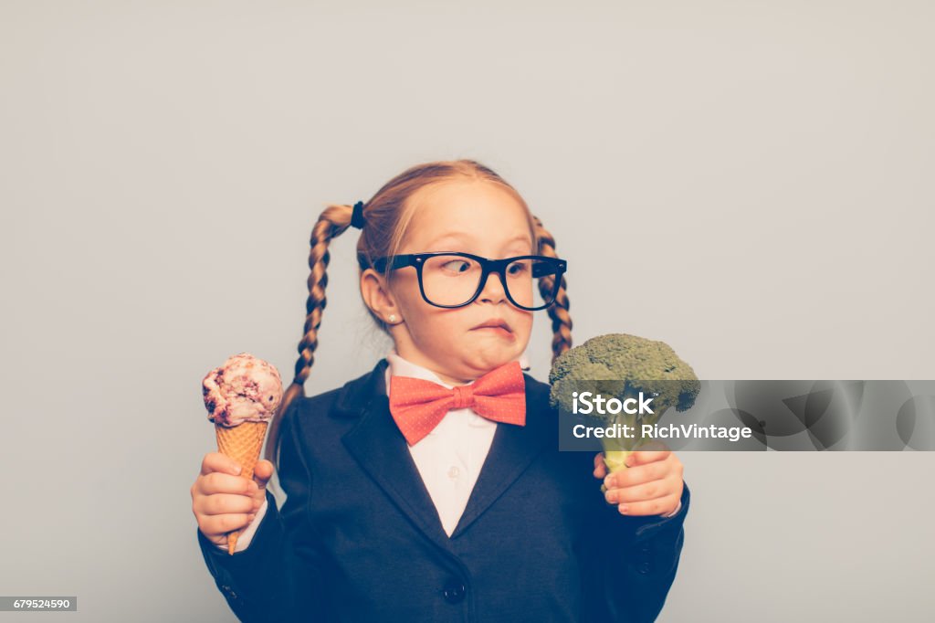 Young Female Nerd Holds Ice Cream and Broccoli A young female nerd dressed in bow tie and eyeglasses is deciding between eating an ice cream cone or broccoli. She is making a disgusted face at the broccoli. She is choosing the treat. Choice Stock Photo
