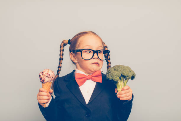 Young Female Nerd Holds Ice Cream and Broccoli A young female nerd dressed in bow tie and eyeglasses is deciding between eating an ice cream cone or broccoli. She is making a disgusted face at the broccoli. She is choosing the treat. sugar food photos stock pictures, royalty-free photos & images