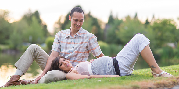 Pregnant Hawaiian couple lounging in grassy park area outside by water