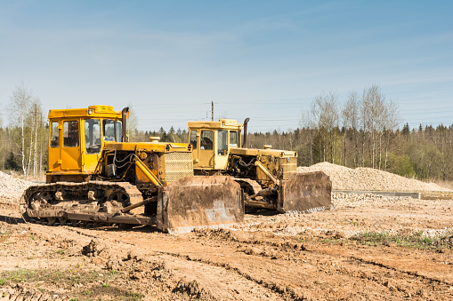 Two yellow dirty old bulldozer stand on a construction site on a sunny day against a background of forest and blue sky