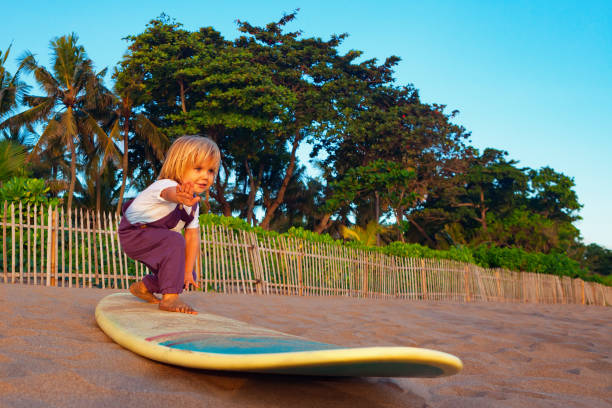 młody surfer stoi na desce surfingowej z zabawą na plaży o zachodzie słońca - one baby boy only zdjęcia i obrazy z banku zdjęć