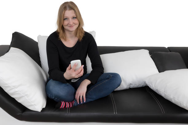 istock Smiling young woman is using her smart phone sitting cross-legged on a black and white couch 679814250