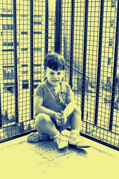 Blue toned vintage photo of a child sitting on the corner of an urban balcony.