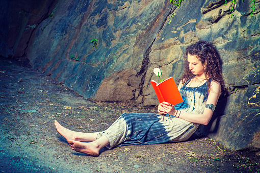 Girl Reading Outside. Wearing long dress, barefoot, a pretty teenage college student with curly long hair is sitting on ground against rocks, holding a red book and a white rose, reading, relaxing.\