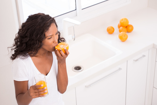 Woman biting half of orange fruit juice, drinking orange juice, standing at kitchen with white sink.