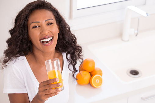 Happy woman drinking fresh orange juice, standing on kitchen with white sink.