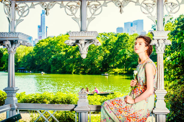 Young Lady Waiting for You. Dressing in sleeveless, long dress, a pretty teenage girl is sitting inside a pavilion, hands holding a white rose, looking at you. people rolling boats in background,.\