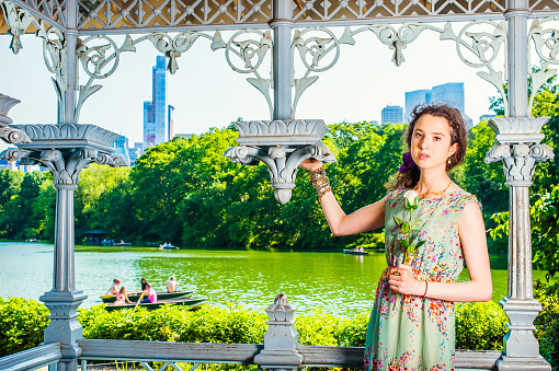 Young Lady Waiting for You. Pretty teenage girl wearing sleeveless, long dress, standing inside pavilion at park, hand holding white rose, looking at you. people rolling boats on background.