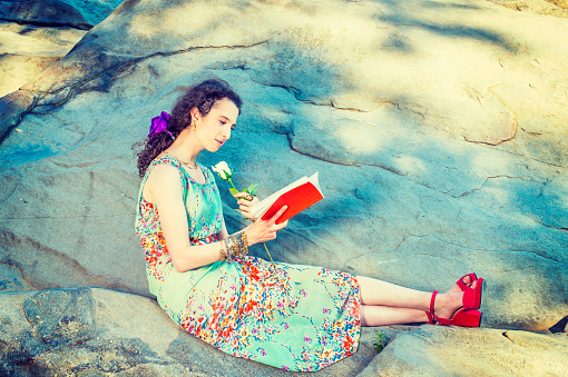 Woman Reading Outside. Beautiful college student wearing sleeveless, long dress, red sandals shoes, chunky chain bracelet, sitting on rocks, hands holding white rose, reading red book, relaxing.