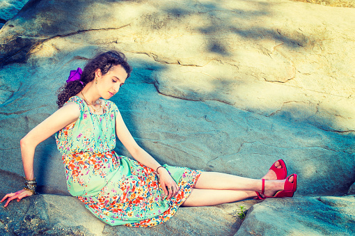 Summer Fashion. Dressing in sleeveless, long dress, red sandals shoes, purple flower hairpin,  chunky chain bracelet, a beautiful teenage girl is sitting on a big rocks outside, relaxing, thinking,\