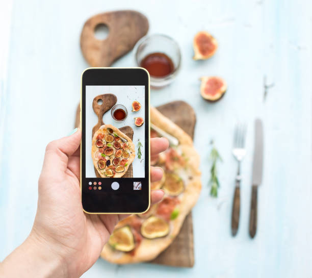 istock Hand making shot on mobile phone: rustic homemade pizza with figs, prosciutto and mozzarella cheese, dark wooden serving board over light blue backdrop. Top view 679891594