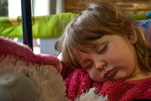 Blond baby girl wearing a pink fur sweater asleep on a chair with a furry teddy bear on her lap.