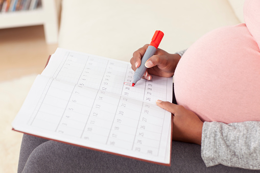 Baby due date - unrecognizable pregnant woman wi big belly in pink t-shirt marking off dates on calendar, using red marker.