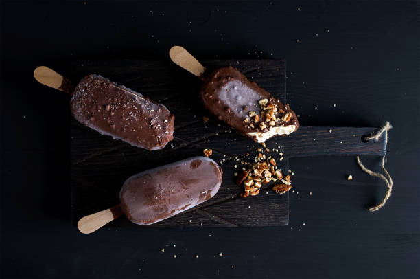 chocolate dipped popsicles with chipped nuts on dark wooden board over black background - milk old fashioned retro revival still life imagens e fotografias de stock