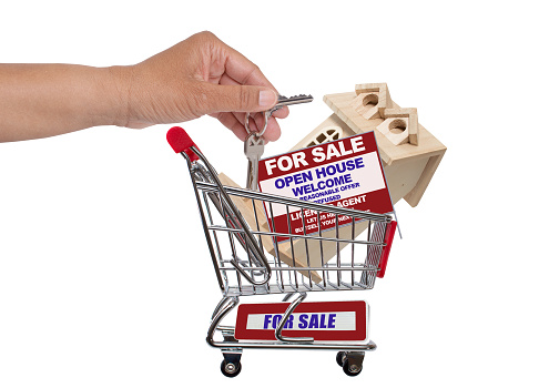 Hand dropping key in shopping cart filled with wood house, real estate for sale open house sign isolated on white background