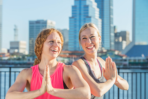 Two women doing yoga outdoors in the city, on the waterfront, on a sunny day. They are standing with palms pressed together in a prayer position.