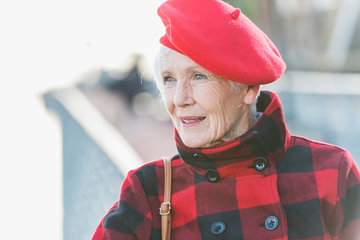 Headshot of a stylish senior woman in her 70s with blue eyes and white hair standing outdoors. She is wearing a red flat cap and red plaid coat.