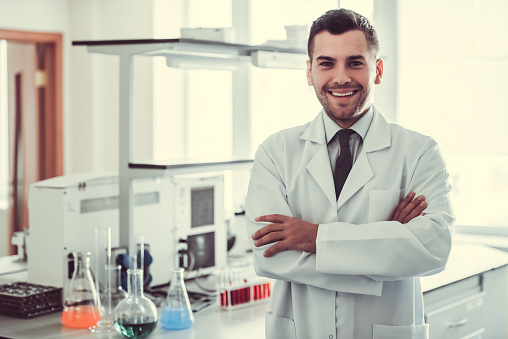 Handsome medical doctor in white coat is looking at camera and smiling while standing in laboratory