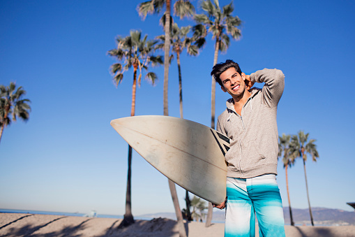Smiling young man with surfboard at Venice Beach. Male is standing while looking away on shore. He is enjoying summer vacation.