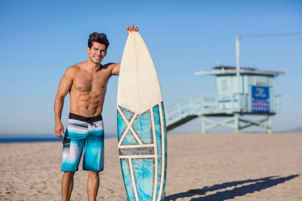 lächelnder junger mann mit surfbrett am venice beach - swim truncks stock-fotos und bilder