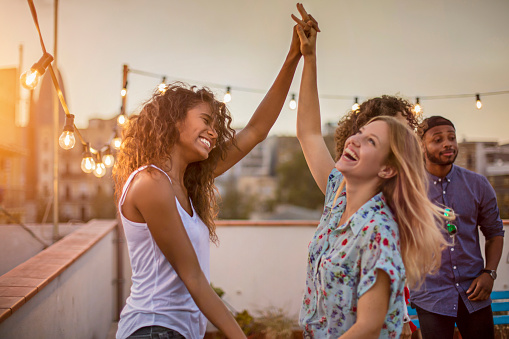 Cheerful women dancing with arms raised during party. Happy female friends are enjoying on terrace. Multi-ethnic people are celebrating during sunset.