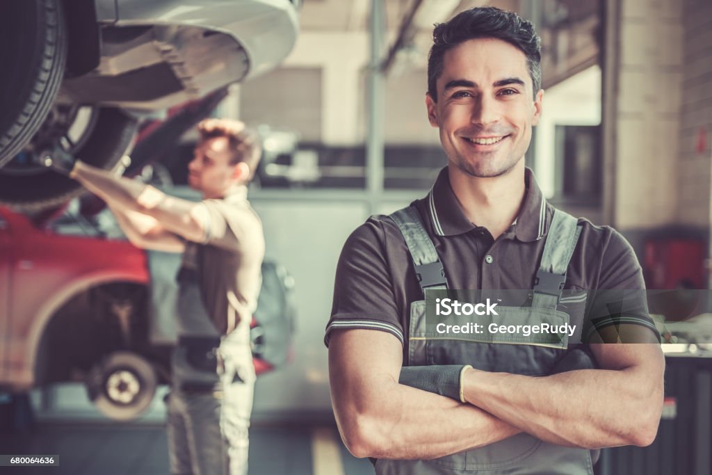 At the auto service At the auto service. Handsome young auto mechanic in uniform is looking at camera and smiling while his colleague is examining car Mechanic Stock Photo